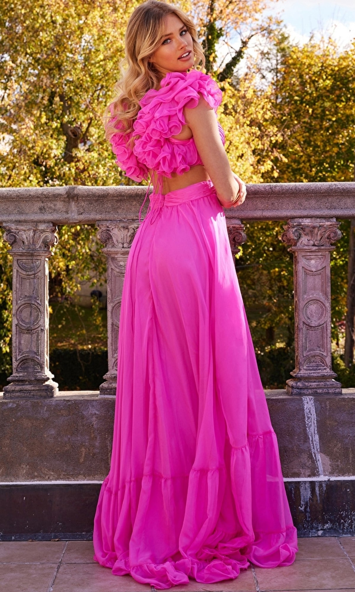 A woman stands outdoors by a stone railing, wearing the Jovani Ruffled-Bodice Long Tiered Prom Dress 23322 in bright pink. The floor-length A-line dress features ruffled sleeves and back, with trees and sunlight in the background.